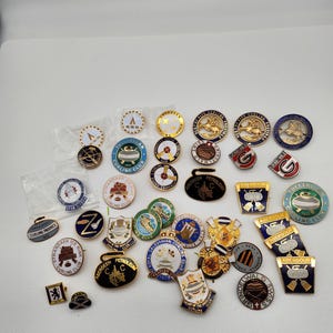 Puede incluir: Una colección de 30 pines redondos de color dorado con varios diseños. Los pines presentan imágenes relacionadas con el curling, incluyendo piedras de curling, pistas de hielo y logotipos de clubes. Algunos pines tienen texto, como "Aberdeen Petroleum Curling Club" y "Tulliallan Curling Club".