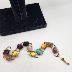 Puede incluir: Un brazalete de tono dorado con un diseño de cadena de eslabones y piedras de varios colores. Las piedras están engastadas en engastes de tono dorado e incluyen amatista, verde, blanco, naranja y rojo.