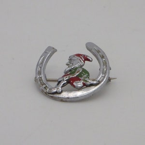 May include: A silver horseshoe brooch with a red and green leprechaun sitting inside. The leprechaun is wearing a red hat and a green shirt.