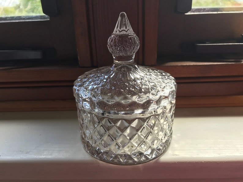 May include: A clear glass jar with a diamond-shaped pattern and a pointed lid. The lid has a decorative design of small circles.