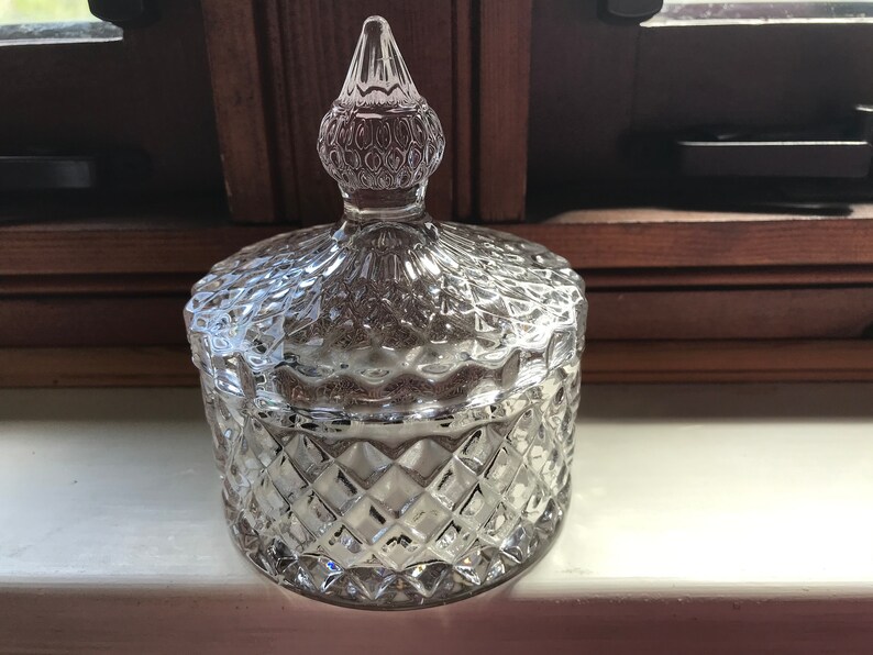 May include: A clear glass jar with a diamond-shaped pattern and a domed lid with a pointed top. The lid is also decorated with a diamond-shaped pattern.