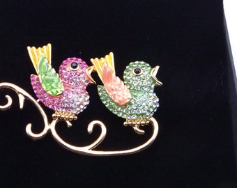 May include: A gold-tone brooch featuring two jeweled birds perched on a decorative branch. The birds are adorned with pink, green, and clear rhinestones.