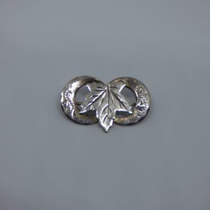May include: Silver brooch featuring a stylised maple leaf design. The leaf is positioned in the centre of a double circle.
