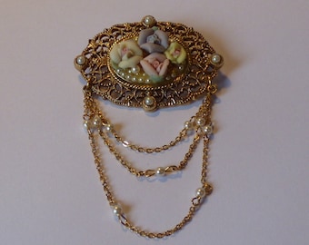 May include: A gold-tone brooch with a floral design featuring pastel pink, purple, and white flowers. The brooch has a dangling chain with multiple strands of pearls.