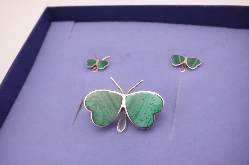 May include: A silver butterfly brooch with green malachite wings. The brooch is displayed in a blue velvet box with two matching earrings.