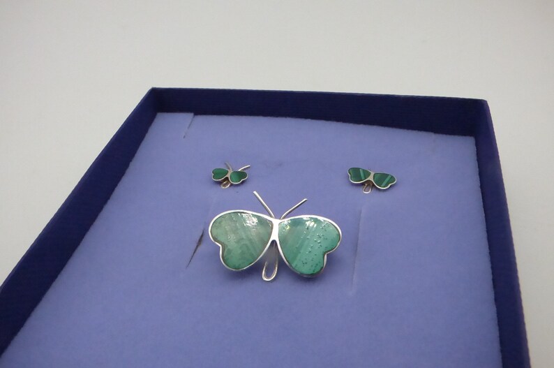 May include: A set of three silver butterfly earrings with green malachite stones. The earrings are displayed in a blue velvet box.