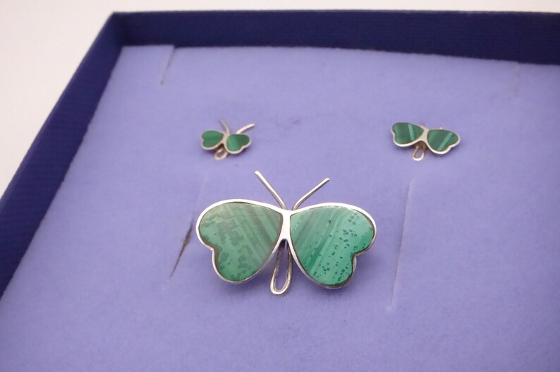 May include: A set of three silver butterfly jewelry pieces with green malachite stone accents. The largest piece is a brooch, and the smaller pieces are earrings.