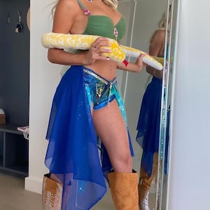May include: A woman wearing a green bra, blue denim shorts, and a blue sheer skirt. She is holding a yellow snake toy and wearing brown suede boots with rhinestones.