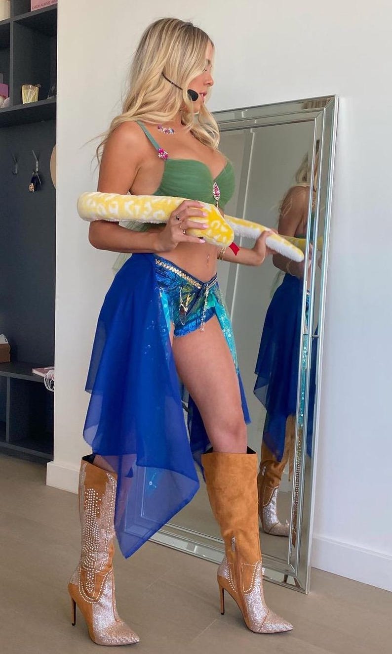 May include: A woman wearing a green bra, blue denim shorts, and a blue sheer skirt. She is holding a yellow snake toy and wearing brown suede boots with rhinestones.