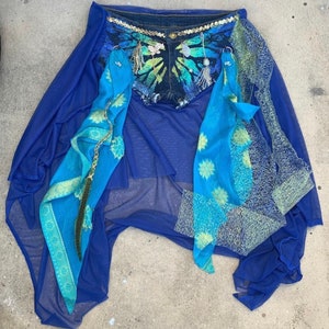 May include: A blue denim skirt with a tie-dye pattern and embellishments. The skirt is layered over a sheer blue fabric with a blue and gold patterned scarf.