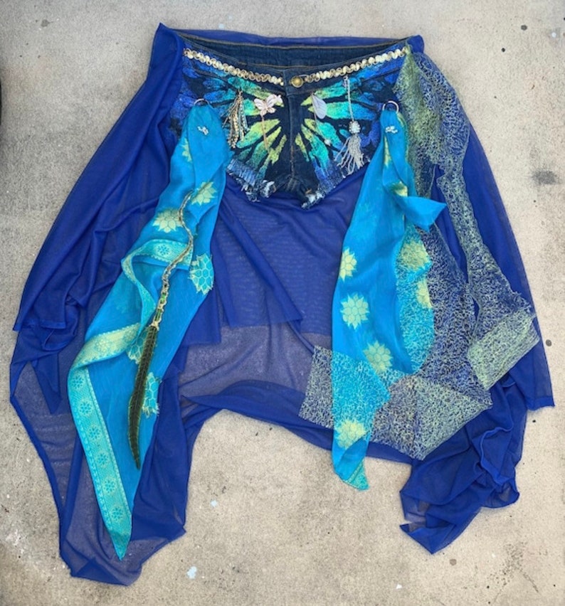 May include: A blue denim skirt with a tie-dye pattern and embellishments. The skirt is layered over a sheer blue fabric with a blue and gold patterned scarf.