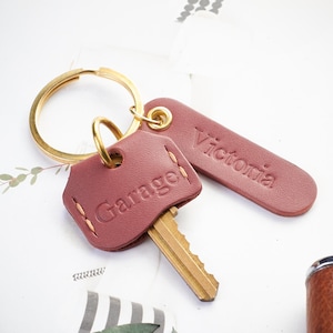 Könnte beinhalten: Ein burgunderfarbenes Leder-Schlüsselanhänger-Set mit einem goldenen Schlüsselring. Das Set enthält einen Schlüsselanhänger mit dem geprägten Schriftzug "Garage" und einen rechteckigen Anhänger mit dem geprägten Schriftzug "Victoria". Ein goldener Schlüssel ist in den Schlüsselanhänger eingesetzt.