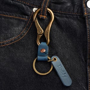 May include: A keychain featuring an antique brass carabiner clip attached to a blue leather strap with a copper rivet. A circular brass ring is connected to the strap, along with a rectangular blue leather tag embossed with the word Daddy.