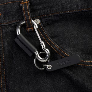 Könnte beinhalten: Ein silberfarbener Karabiner-Schlüsselanhänger mit einer schwarzen Lederhülle am Clip und einem schwarzen, rechteckigen Lederanhänger, in den der Name David eingraviert ist. Der Schlüsselanhänger ist an einem silbernen Metallring befestigt.