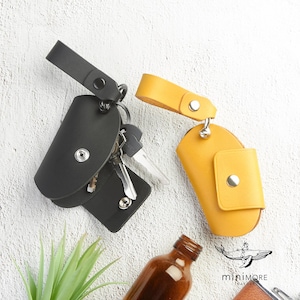 May include: Two leather key holders, one black and one yellow, are displayed against a white background. The black key holder is open, revealing keys. The yellow key holder is closed. Both have a snap closure and a wrist strap.