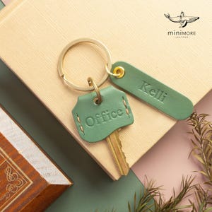 May include: A sage green leather key cover embossed with the word Office, attached to a gold-tone metal keyring along with a matching rectangular leather tag embossed with the name Kelli. The key cover features visible stitching along the edges.