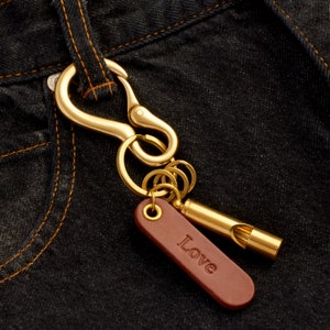 May include: A gold-toned key chain with a brass whistle and a leather tag that says "Love". The keychain is hanging on a pair of blue denim jeans.