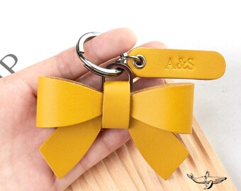 May include: A mustard yellow leather bow keyring featuring a silver metal split ring and a rectangular leather tag embossed with the letters A&S. The bow is constructed from folded leather strips secured by a central leather band.