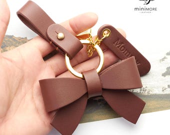 Peut inclure: Un porte-clés en cuir marron avec un grand nœud, un anneau en métal doré et une petite étiquette rectangulaire en cuir gravée du mot Mommy. Le porte-clés comprend une lanière avec un bouton-pression doré pour la fixation.