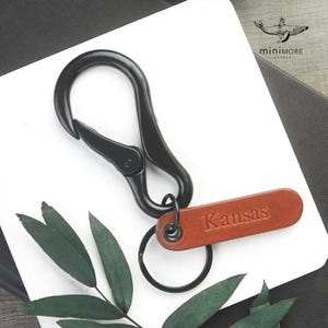 Può includere: Un portachiavi con moschettone in metallo nero dotato di un'etichetta rettangolare in pelle marrone con impresso il nome Kansas. L'etichetta in pelle è fissata al moschettone tramite un piccolo anello in metallo nero. Logo con balena.
