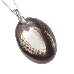 May include: Silver chain necklace with an oval pendant. The pendant features a clear, oval-shaped glass or resin encasing strands of dark brown hair. The pendant is approximately 1.5 inches long.