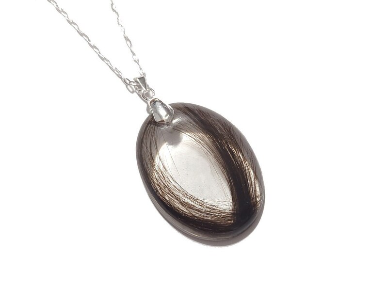 May include: Silver chain necklace with an oval pendant. The pendant features a clear, oval-shaped glass or resin encasing strands of dark brown hair. The pendant is approximately 1.5 inches long.