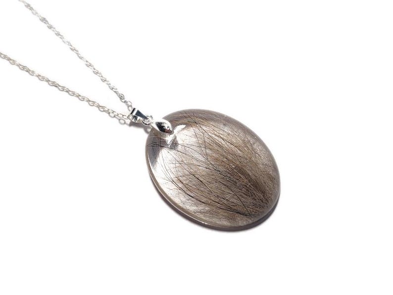 May include: Silver pendant necklace with an oval-shaped, clear resin pendant containing strands of gray hair. The pendant is suspended from a delicate silver chain. The background is plain white.