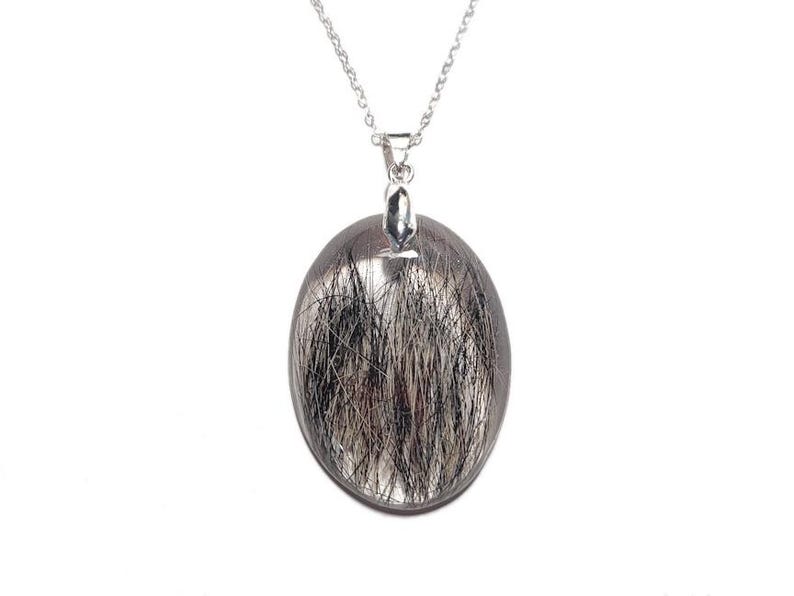 May include: A silver necklace featuring an oval pendant with a clear, transparent appearance. The pendant is filled with black and silver needle-like inclusions, creating a unique, textured effect. The chain is delicate and silver.