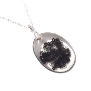 May include: A silver necklace featuring a clear, oval pendant encasing a cluster of dark, curly material. The pendant is suspended from a delicate silver chain, creating a minimalist and elegant design. The necklace is set against a white background.