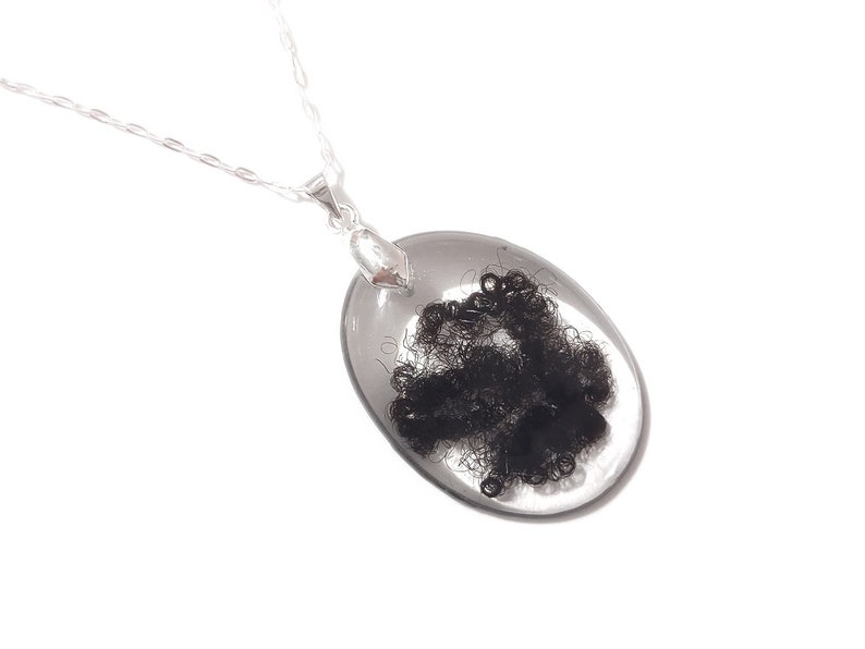 May include: A silver necklace featuring a clear, oval pendant encasing a cluster of dark, curly material. The pendant is suspended from a delicate silver chain, creating a minimalist and elegant design. The necklace is set against a white background.
