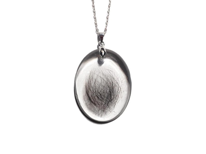 May include: A silver pendant necklace with an oval, clear glass or resin pendant. The pendant contains a small bundle of dark hair. The necklace chain is also silver and hangs against a white background.