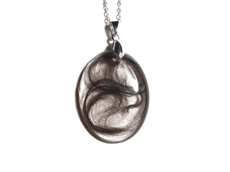 May include: Oval-shaped pendant necklace with dark hair encased in clear resin. The pendant hangs from a silver chain and clasp, set against a white background. The hair appears to be dark brown or black.