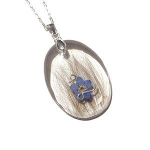May include: Silver-toned necklace featuring an oval pendant with a clear resin encasing. Inside the resin are strands of hair and a small blue forget-me-not flower. The pendant is suspended from a delicate chain.