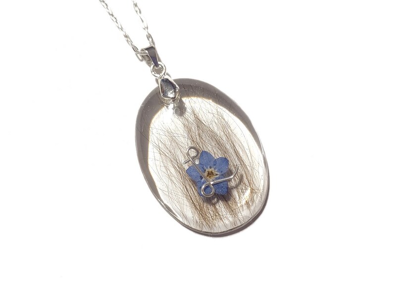 May include: Silver-toned necklace featuring an oval pendant with a clear resin encasing. Inside the resin are strands of hair and a small blue forget-me-not flower. The pendant is suspended from a delicate chain.