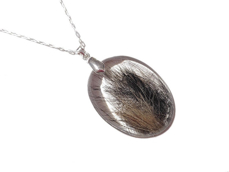 May include: Oval pendant necklace with a clear resin stone, featuring black and brown inclusions. The pendant is suspended from a delicate silver chain. The necklace is displayed against a white background, highlighting the pendant's unique design.