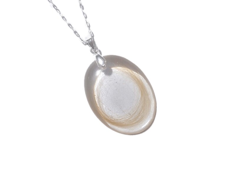 May include: A silver necklace with a clear, oval pendant. The pendant contains a swirl of light brown hair. The chain is delicate and the pendant is smooth and polished.