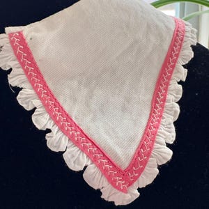 May include: White, triangular handkerchief with a pink stitched border and ruffled edges. The handkerchief is made of a soft fabric and is displayed on a black surface. The pink border has a decorative stitch pattern.