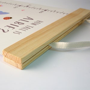 May include: A wooden growth chart with a white background and a light brown wooden frame. The chart has a white ribbon attached to the frame. The text "How Tall Is Albie?" is printed on the chart.