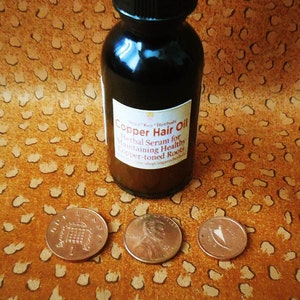 May include: A dark brown glass bottle with a dropper top and a label that reads "Copper Hair Oil Herbal Serum for Maintaining Healthy Copper-toned Roots". The bottle is sitting on a brown patterned surface with three copper-colored coins.