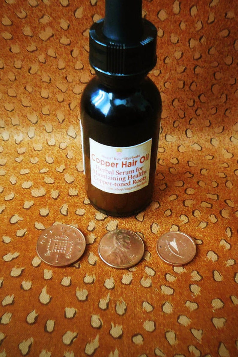 May include: A dark brown glass bottle with a dropper top and a label that reads "Copper Hair Oil Herbal Serum for Maintaining Healthy Copper-toned Roots". The bottle is sitting on a brown patterned surface with three copper-colored coins.