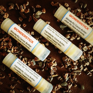 May include: Four white tubes of cocoa butter lip balm with a brown label that reads "WONDERSTICK" and "Wonderful Cocoa Butter stick for face and body". The tubes are surrounded by cocoa nibs on a brown surface.
