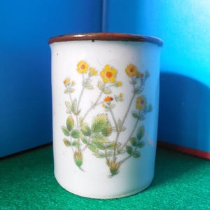May include: White ceramic mug with a brown rim, decorated with a floral design featuring yellow flowers and green leaves. The mug is cylindrical and appears to be a standard size for beverages.