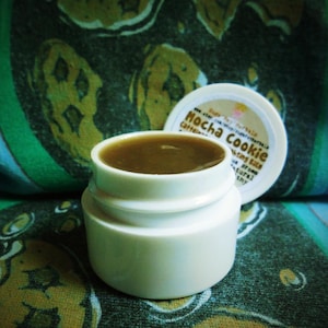 Puede incluir: Un pequeño tarro blanco con un bálsamo labial marrón en su interior. El tarro tiene una etiqueta que dice "Mocha Cookie" y "Bálsamo labial con cafeína".
