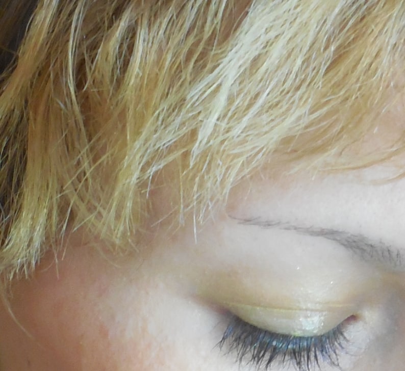May include: Close-up of a person's eye with light blonde hair. The eye is closed and has a light yellow eyeshadow applied.