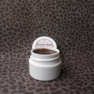 May include: A small white jar with a brown lid containing a dark brown balm. The lid has the text "Sugar-Rey Herbals Brownie Balm Encourages healthy hair" printed on it.
