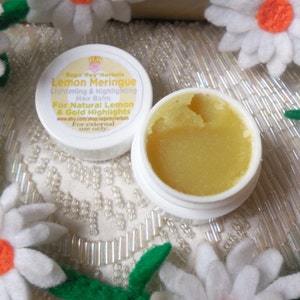 May include: A white jar of Lemon Meringue Hair Balm by Suga Rey Herbals. The jar is open and the balm is a light yellow color. The jar is surrounded by white felt flowers.