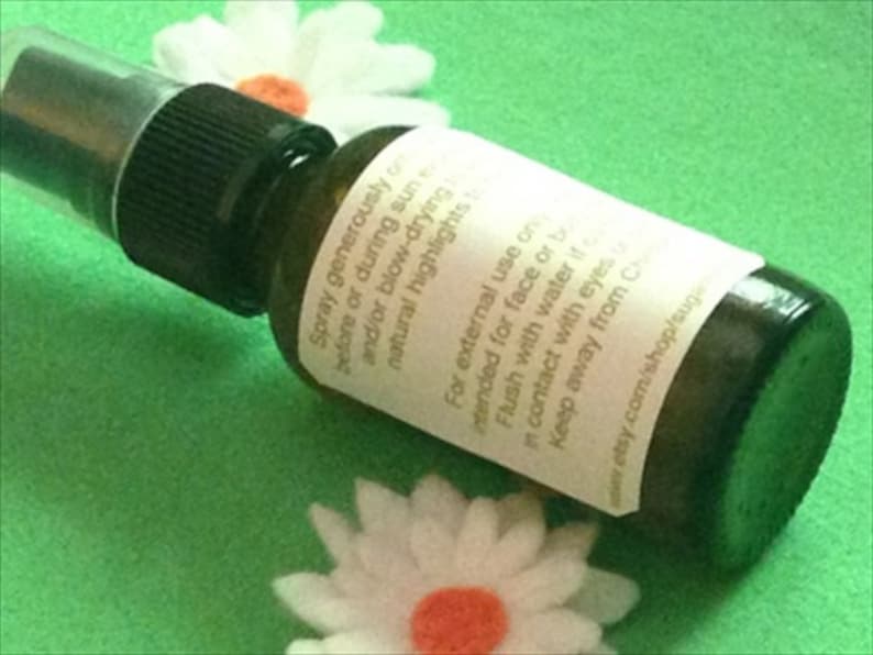 May include: A clear glass bottle with a black spray nozzle and a white label. The label has text that reads "Spray generously before or during sun and/or blow-drying natural highlights" and "For external use on intended for face or body. Flush with water if in contact with eyes. Keep away from children. www.etsy.com/shop/sugarplum",  The bottle is sitting on a green surface.