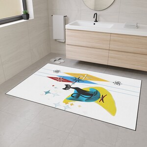May include: A white bath mat with a black cat sitting on a blue and yellow crescent moon. The cat is surrounded by stars and atomic symbols. The mat has a mid-century modern design.