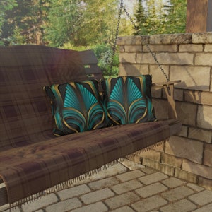 May include: Two teal and gold art deco-style throw pillows rest on a brown plaid swing hanging on a porch. The pillows feature a repeating geometric pattern of stylized leaves. The swing is made of dark brown wood with a brown plaid throw blanket draped over it. The swing hangs from a chain attached to a rustic stone wall.