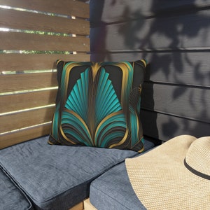 May include: Teal and gold art deco patterned throw pillow. The pillow features a geometric design with teal and gold accents on a black background.  The pillow is shown on a dark blue outdoor couch with a beige sunhat resting on the armrest. The pillow is square-shaped and appears to be made of a soft, durable fabric suitable for outdoor use. The design is reminiscent of 1920s and 1930s art deco style. The pillow would add a touch of elegance and sophistication to any outdoor space.